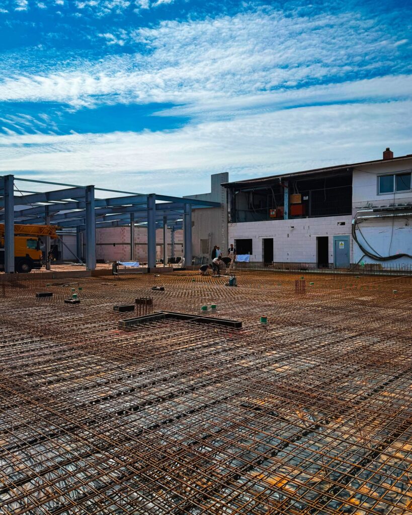 Übersicht Baustelle Bodenplatte: Weitwinkelaufnahme einer großflächigen Stahlbewehrung für eine Bodenplatte, im Hintergrund Arbeiter und ein Autokran.