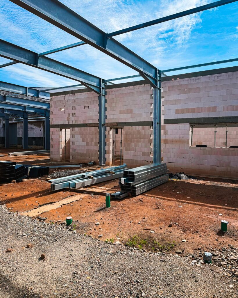 Materiallager auf der Baustelle: Blick in den Rohbau einer Industriehalle mit gestapelten Metallprofilen und Stahlträgern auf rotem Erdboden.