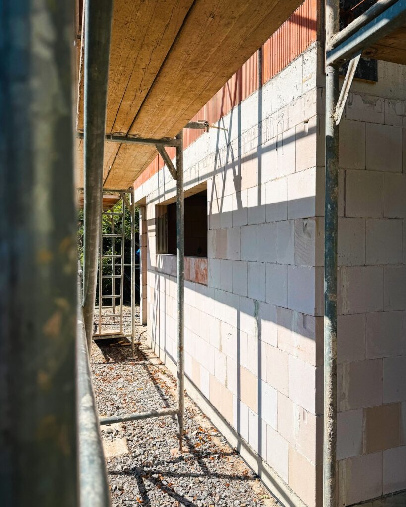 Blick durch ein Baugerüst auf eine frisch gemauerte, helle Wand mit einer Fensteröffnung im Rohbau.