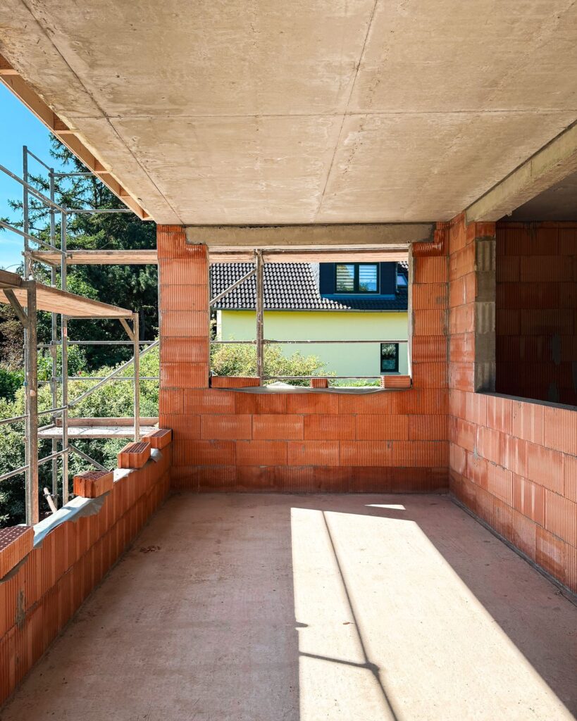 Unfertiger Raum im Obergeschoss mit Sichtbetondecke, rotem Ziegelmauerwerk und einer großen Fensteröffnung mit Blick ins Grüne.