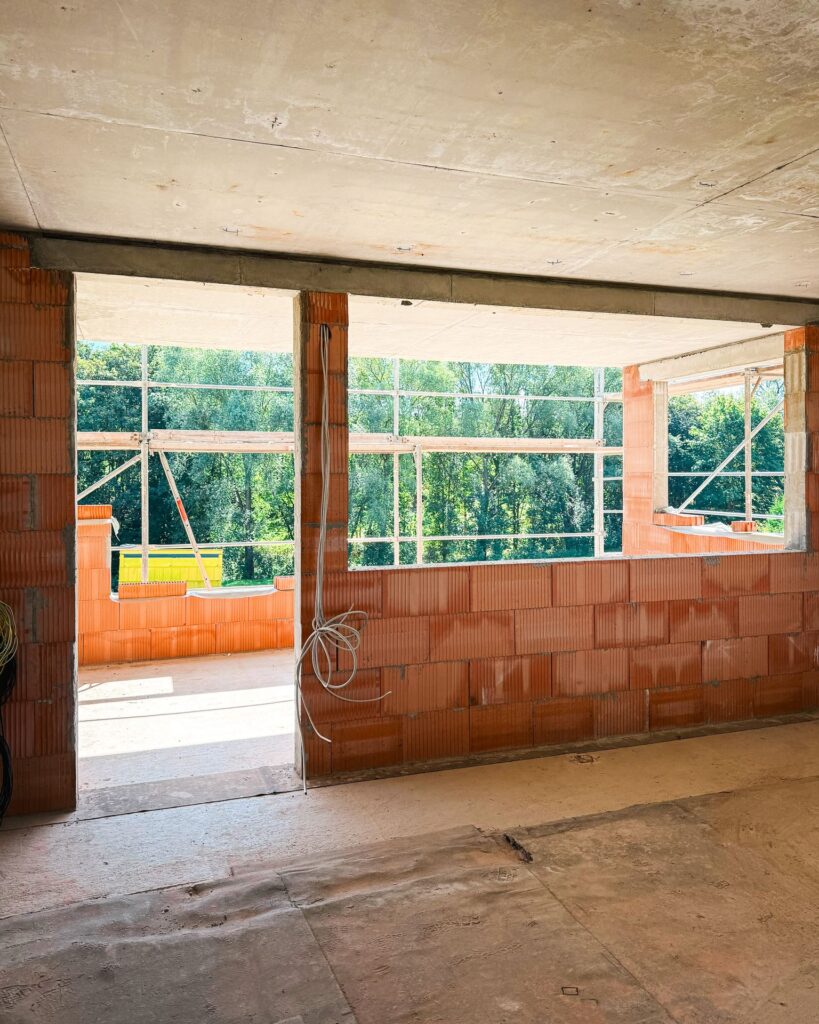 Blick in ein noch offenes Zimmer im Rohbau mit Betonfertigteil-Decke, Ziegelwänden und herabhängenden Kabeln.