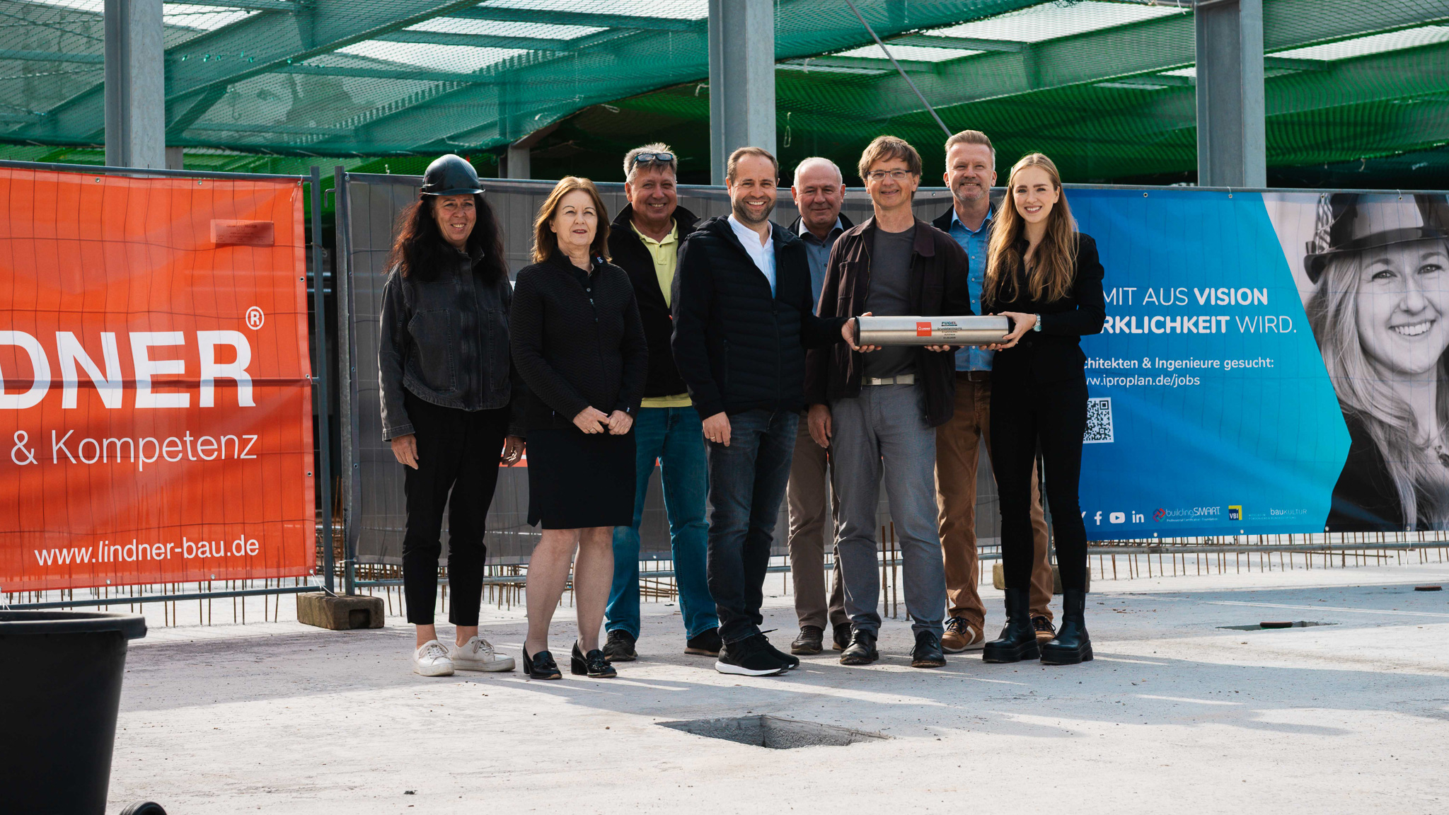Die Projektgruppe bei der Grundsteinlegung für das neue Autohaus Fugel. Acht Personen posieren stolz mit der Zeitkapsel-Hülse auf der Baustelle, im Hintergrund sind Bauzaun-Banner der Firmen Lindner und Fugel zu sehen.