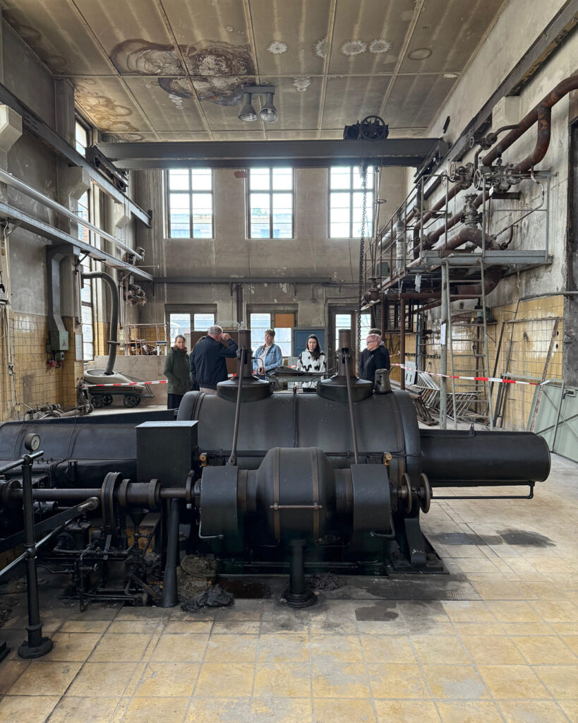 Besucher betrachten eine massive historische Industrie-Dampfmaschine in der Werks halle des ROGO-Werks