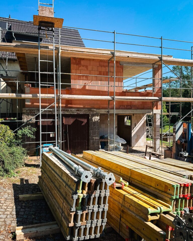 Blick auf einen zweistöckigen Hausanbau im Rohbau mit rotem Ziegelmauerwerk und Baugerüst. Im Vordergrund lagern gestapelte gelbe Schalungsträger und Metallstützen auf einem gepflasterten Weg, links steht ein Lastenaufzug am Gerüst.
