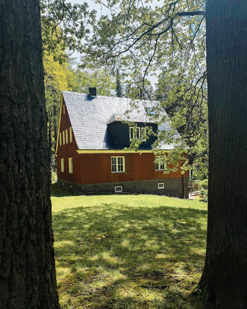 Rustikales rotbraunes Haus mit Schieferdach, eingerahmt von Baumstämmen auf einer sonnigen grünen Wiese im Wald.