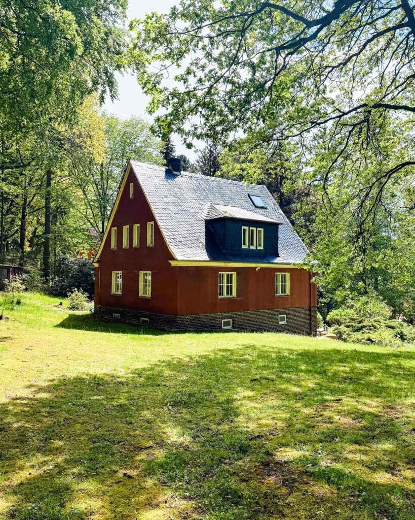 Weitwinkelaufnahme des historischen rotbraunen Wohnhauses mit Schieferdach, eingebettet in einen sonnigen Laubwald.