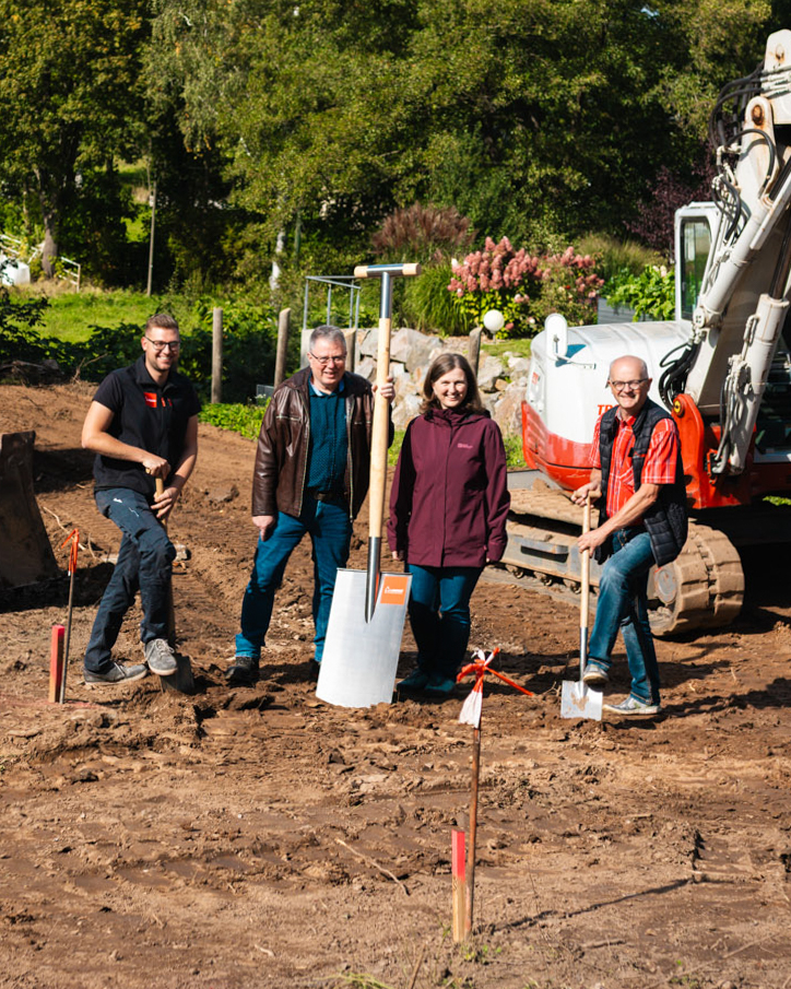 Vier Personen beim Spatenstich auf einer Baustelle mit Bagger im Hintergrund.