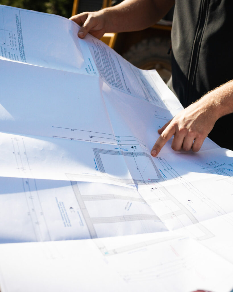 Eine Person in Arbeitskleidung zeigt mit dem Finger auf einen detaillierten Bauplan für den Hausbau.