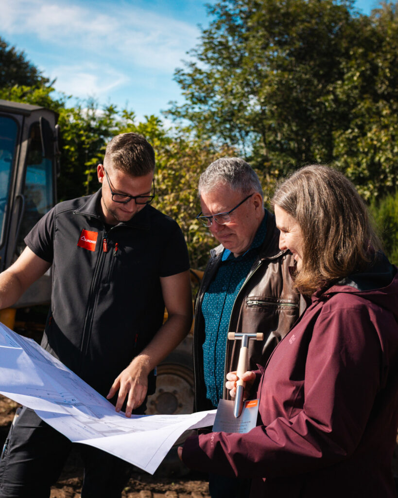 Bauherr, Bauherrin und Bauleiter in Arbeitskleidung besprechen gemeinsam die Baupläne auf der Baustelle.