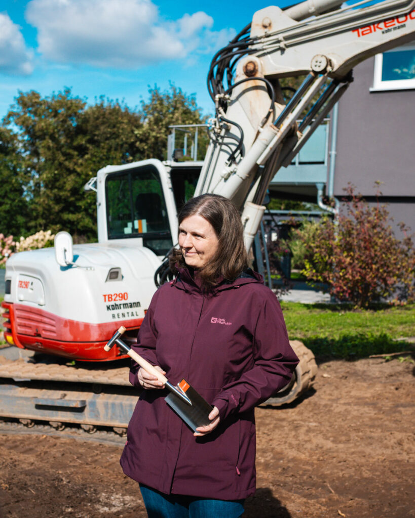 Eine Frau in einer weinroten Jacke hält einen kleinen Spaten vor einem Minibagger auf der Baustelle.