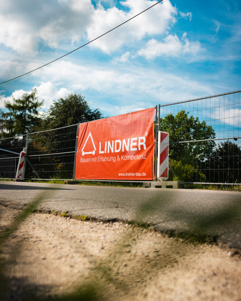 Ein roter Bauzaunbanner der Baufirma Lindner mit dem Slogan "Bauen mit Erfahrung & Kompetenz" und Logo.