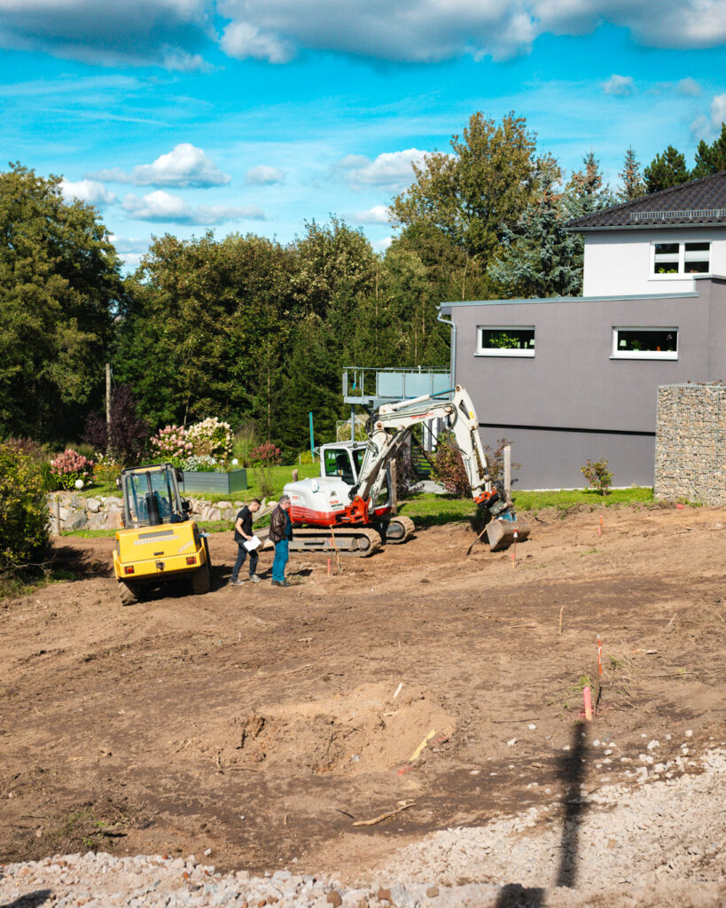 Weitwinkelaufnahme einer Baustelle mit Bagger und Walze vor einem modernen Hausneubau.