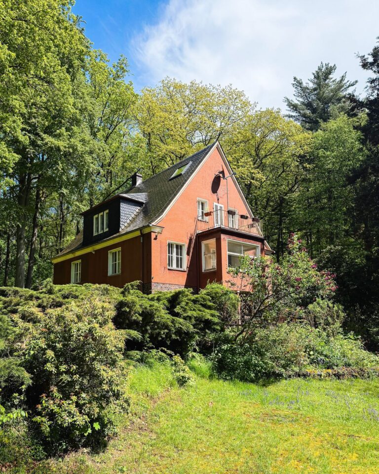 Nahaufnahme des rötlichen Landhauses im Wald mit Balkon, Erker und blühender Vegetation im Garten.