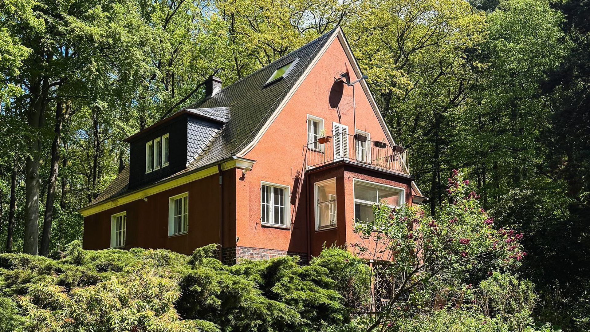 Nahaufnahme des rötlichen Landhauses im Wald mit Balkon, Erker und blühender Vegetation im Garten.